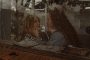 Two young women share an intimate conversation through a cafe window, smiling at each other in warm light.