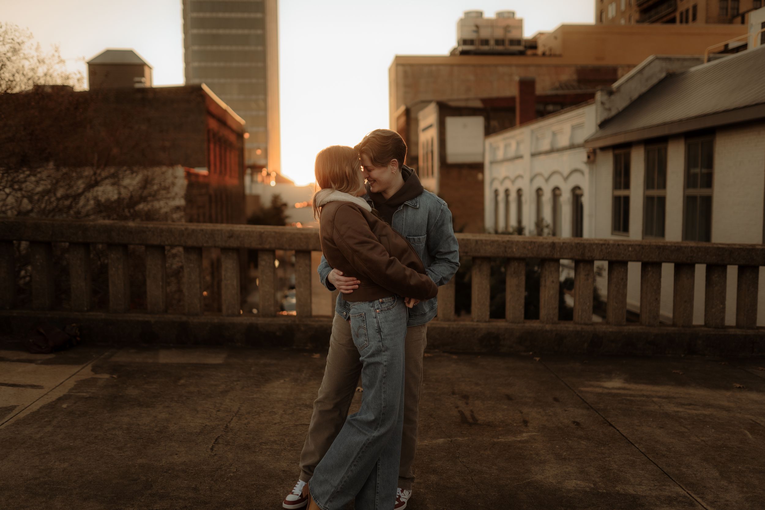 A couple embraces during sunset in a rustic setting with wooden buildings and warm golden light.