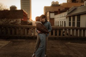 A couple embraces during sunset in a rustic setting with wooden buildings and warm golden light.