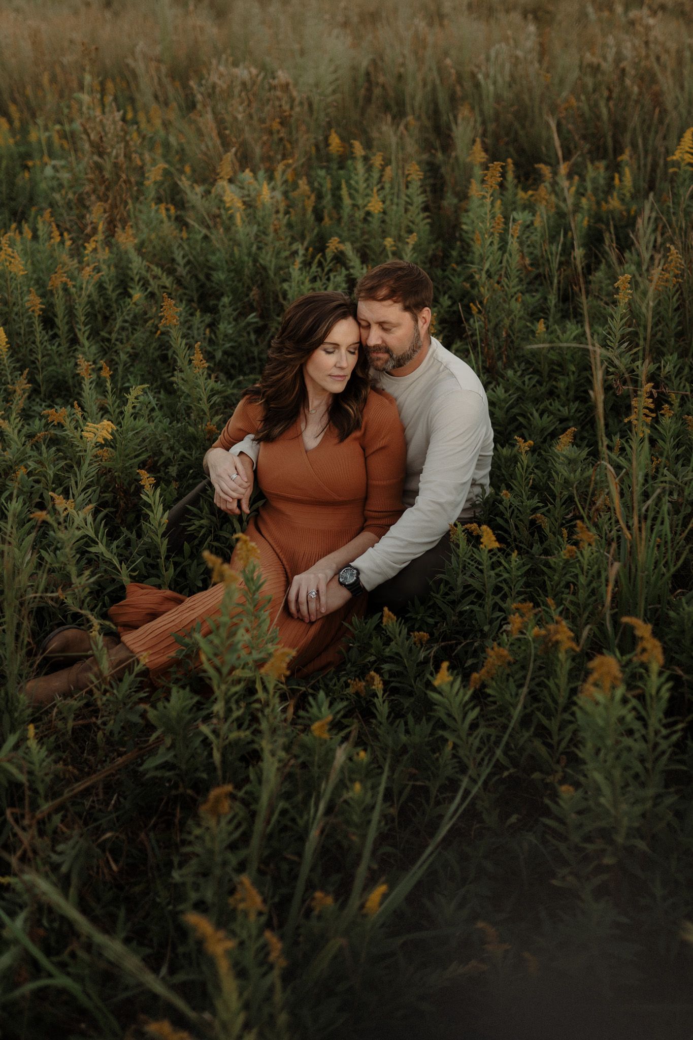A couple sits intimately among tall wildflowers in a golden meadow at sunset.