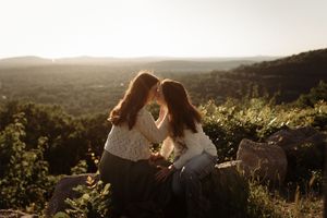 Two women with red hair sit forehead-to-forehead on a rocky hillside at golden hour with scenic valley views.