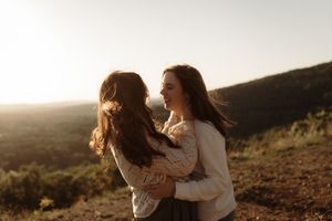 Two young women embracing and smiling at golden hour on a scenic hilltop with rolling green mountains.