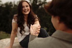 A smiling woman with tattoos holds hands with a person while sitting outdoors, showcasing an engagement ring.
