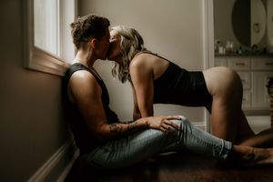 Couple sharing a romantic kiss by a window, man with tattoos in jeans, woman in black top, soft natural lighting.