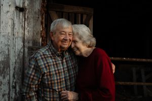 An elderly couple shares a tender moment outside a rustic wooden building.