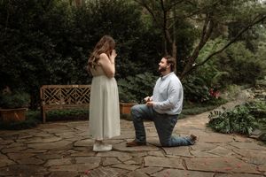 Man kneeling on stone patio proposing to woman in white dress surrounded by lush garden greenery and wooden bench.