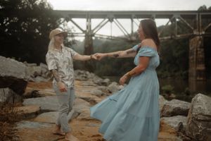 Couple dancing on rocky riverbank near a bridge, woman in blue off-shoulder dress, man in floral shirt and hat.