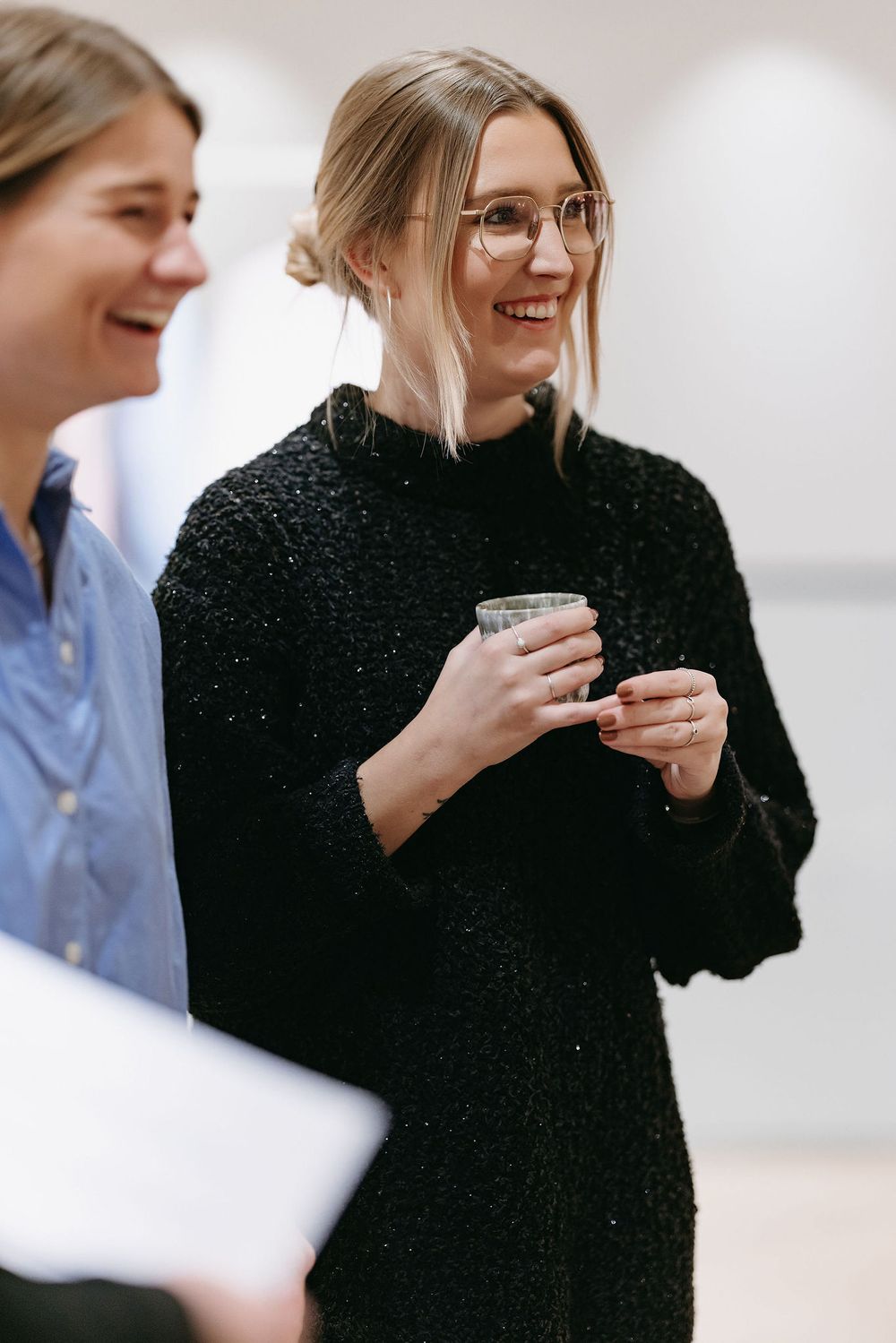 Person in black sweater holds beverage while sharing conversation at indoor event.