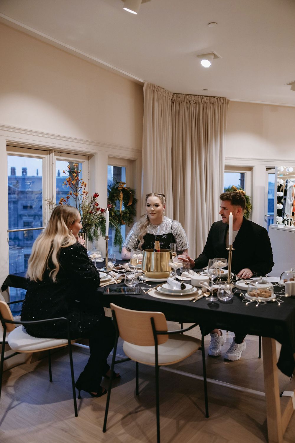 People gather around elegant dinner table setting with city views through windows.