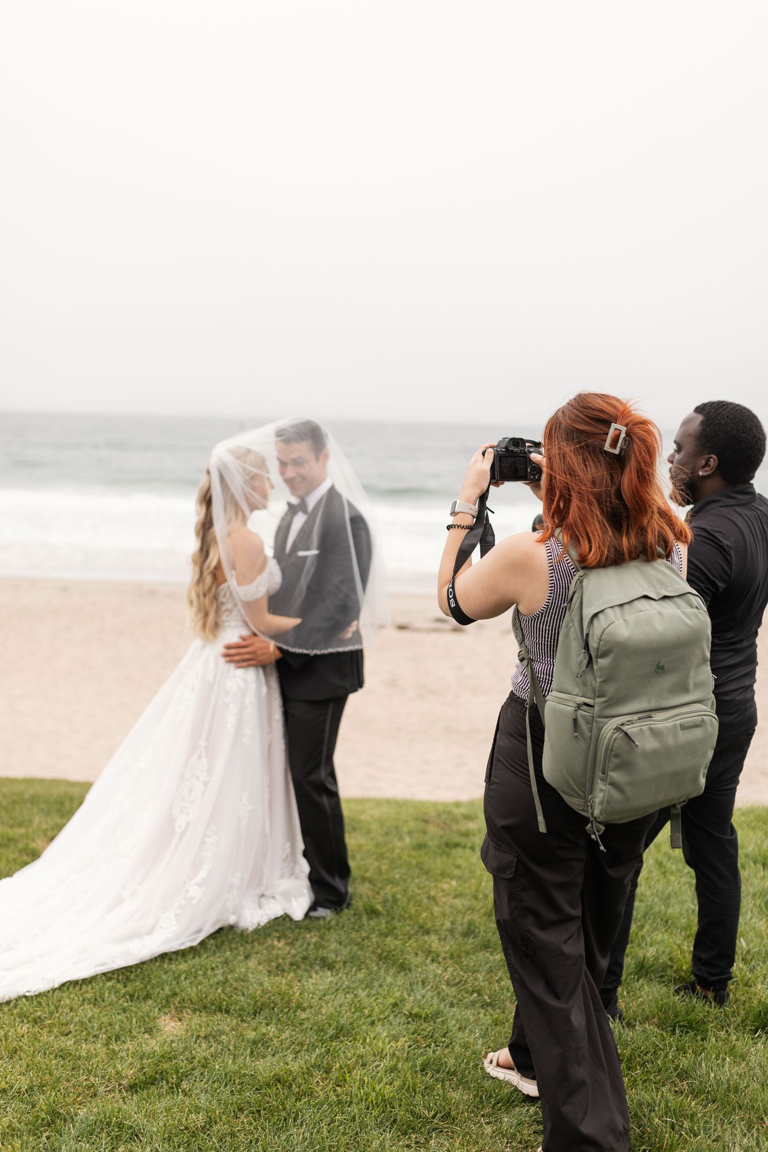 Wedding photographer capturing couple on grassy field near beach.