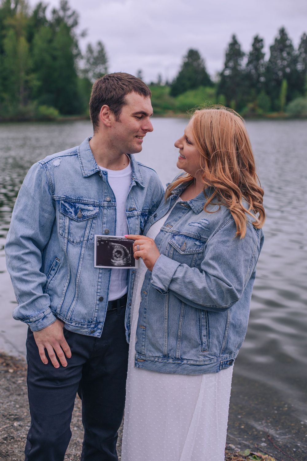 A couple in matching denim jackets share a tender moment by a lake while holding an ultrasound photo. Spokane Liberty Lake Photographer