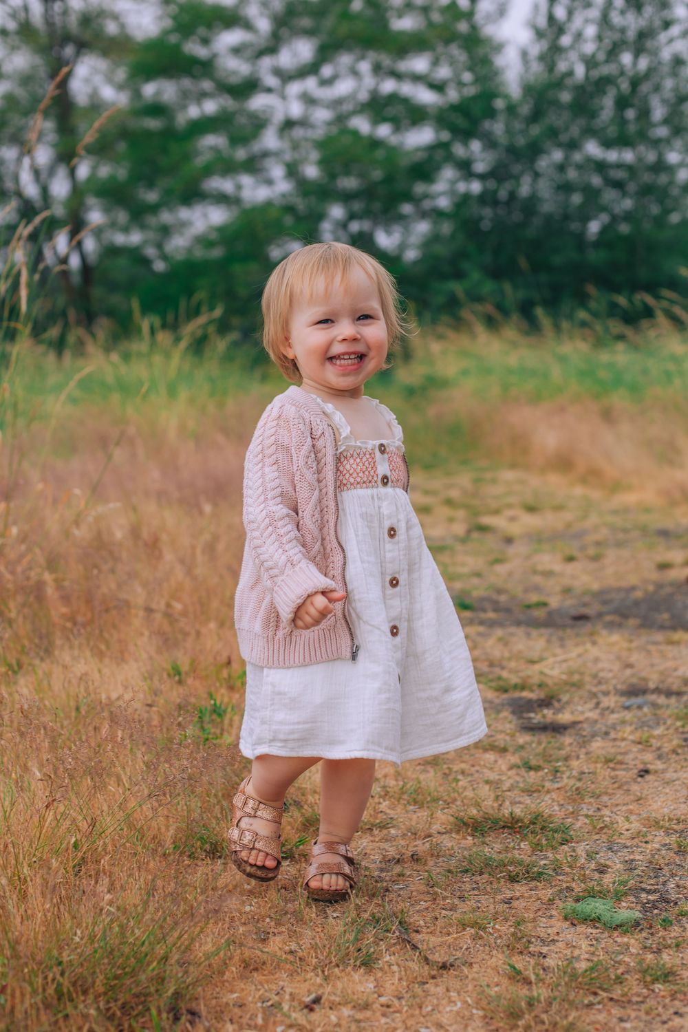 A toddler in a white dress and pink cardigan smiles while standing on a dirt path. Spokane Liberty Lake Photographer Family Photographer