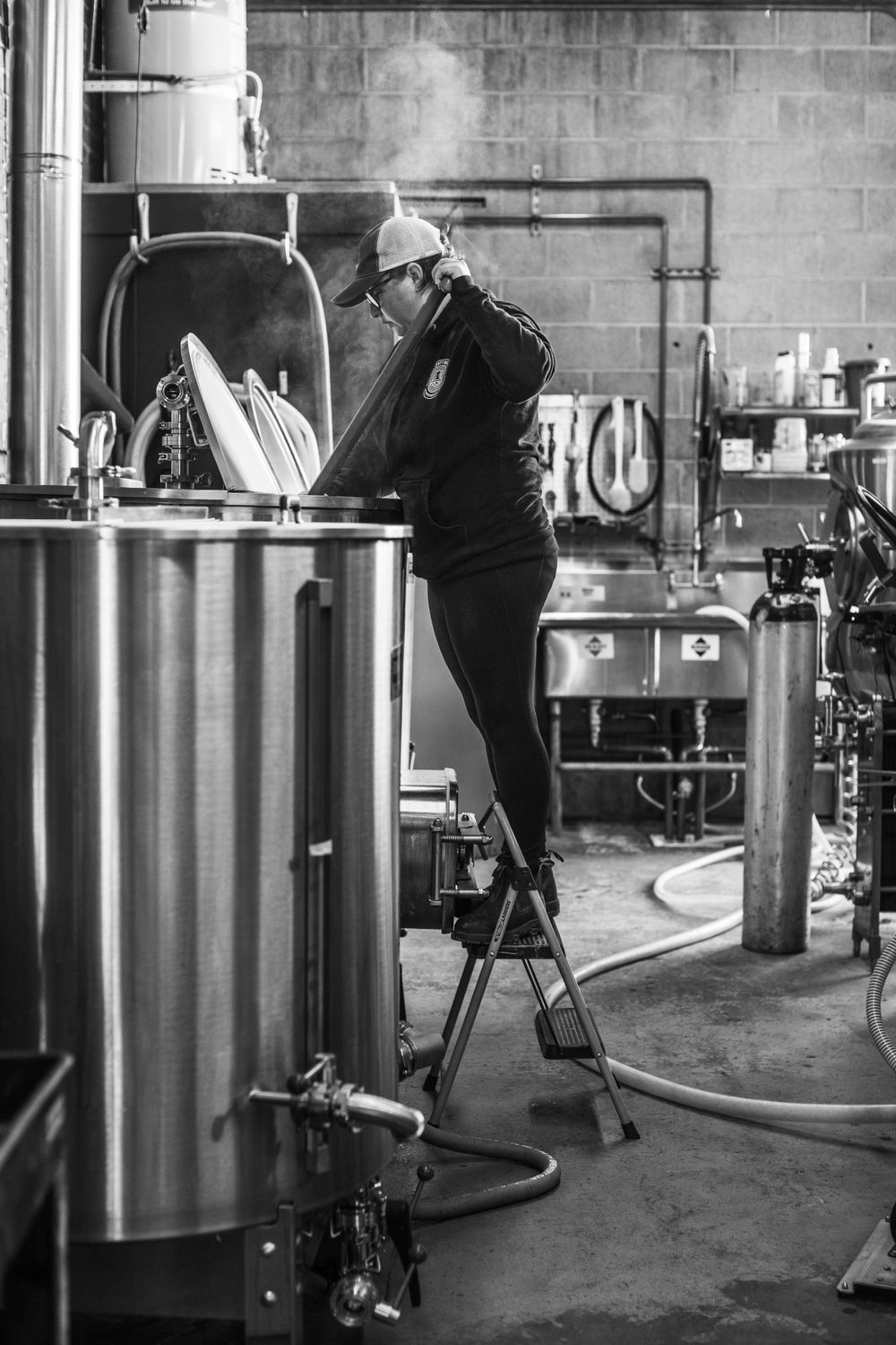 Brewer checking fermentation tanks at craft brewery with ladder and safety equipment.