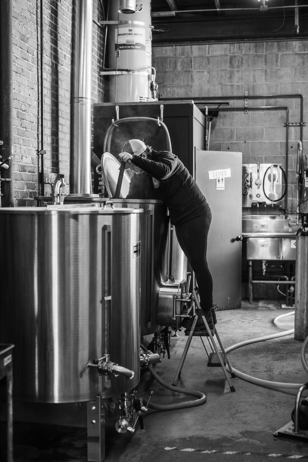 Worker inspecting large stainless steel brewing equipment from elevated position in industrial setting.