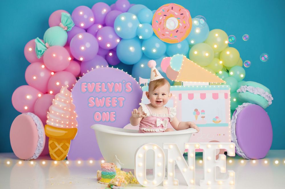 Whimsical sweets-themed birthday party setup with ice cream cones, candy decorations, pastel balloons, and pink lighting. first birthday cak