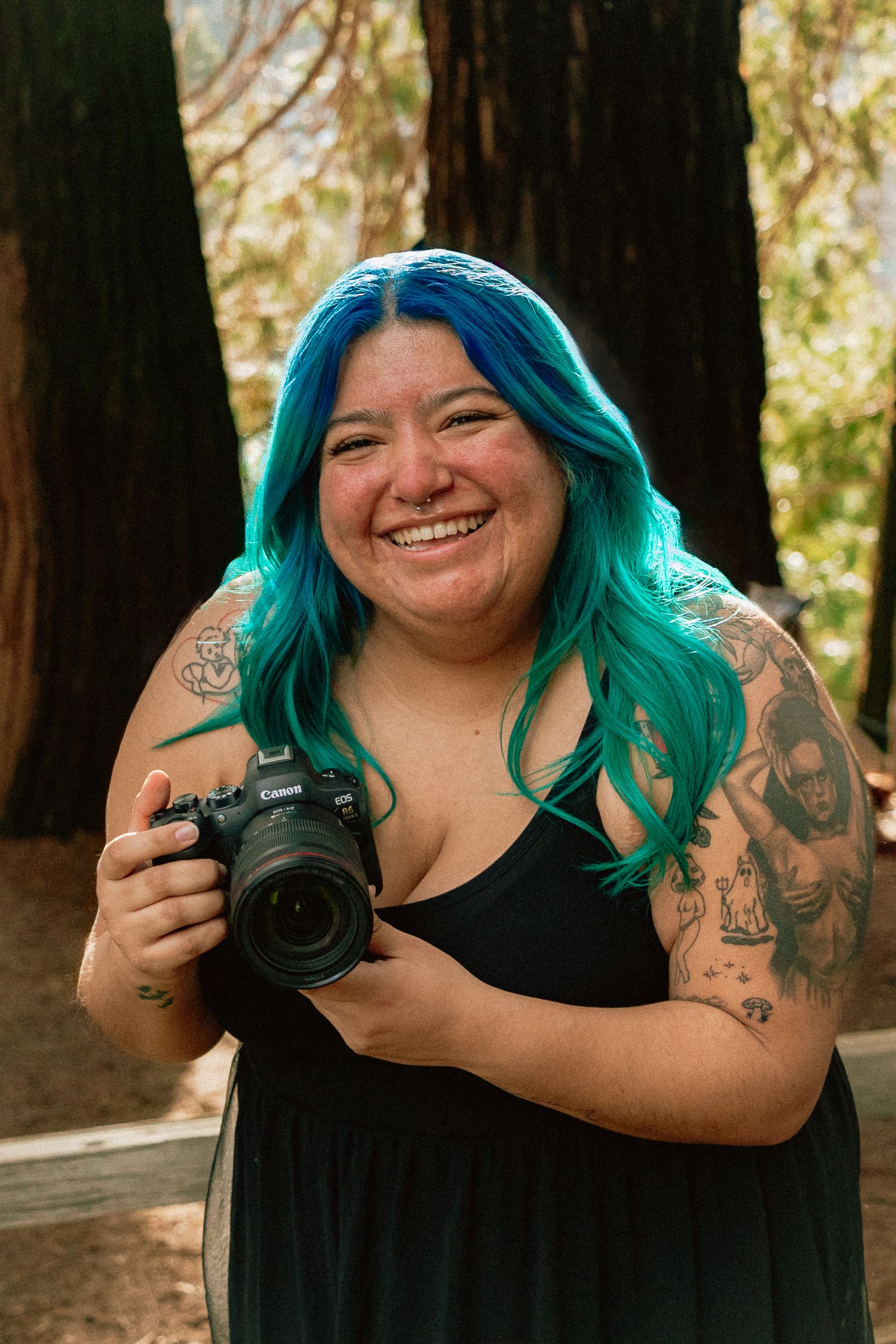 Mexican woman Photographer with bright turquoise hair and tattoos holds professional camera while smiling in forest setting.