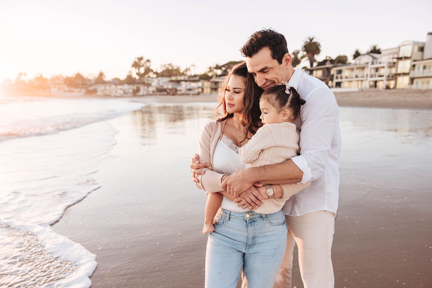 A family embraces with a toddler on dad's shoulders on Miramar Beach, CA during sunset with waves and palm trees in the background.
