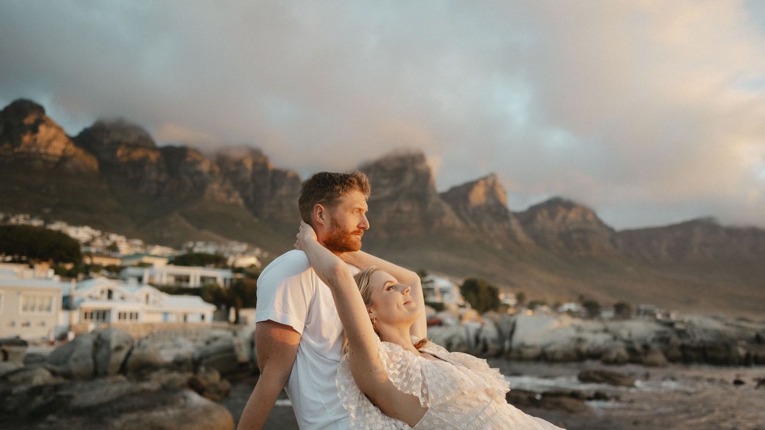 Couple embraces at sunset with coastal mountains and ocean in background.