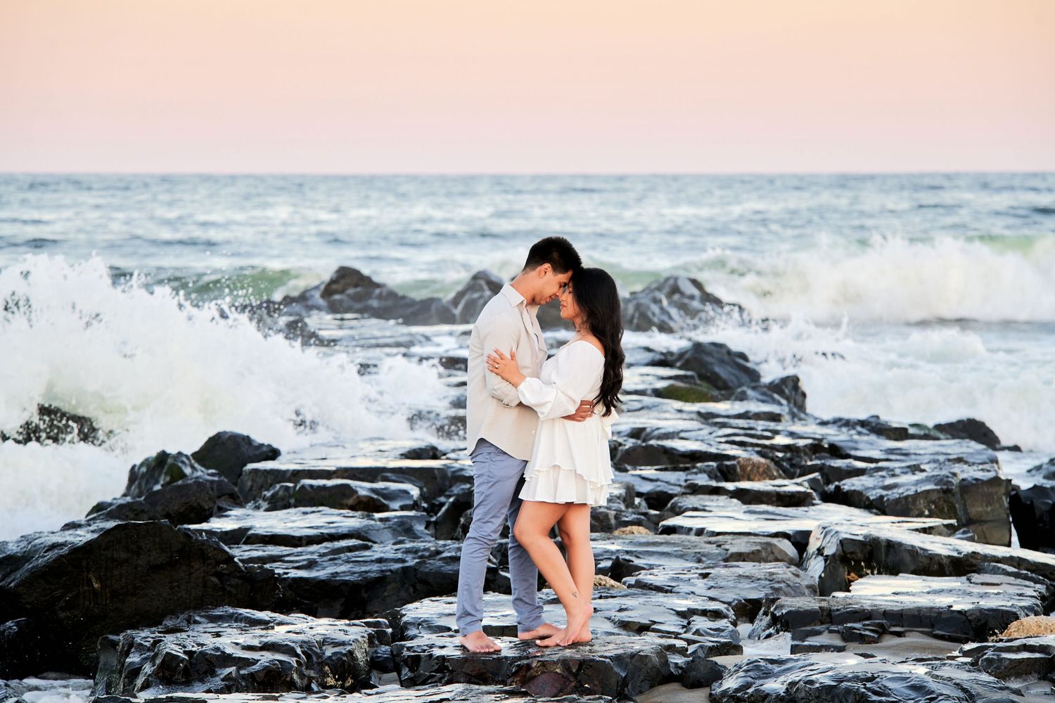 A couple embraces on rocky shore with crashing waves and pink sunset sky in Asbury Park. Gricel's Photography NJ Wedding Photographer