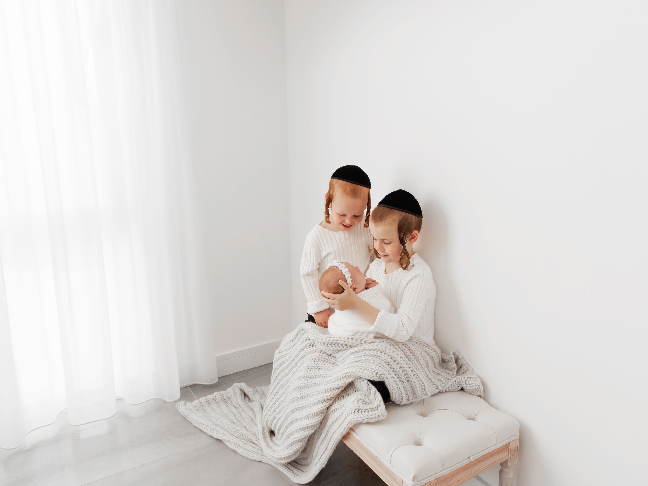 Family snuggling together on white bench near window with soft lighting.
