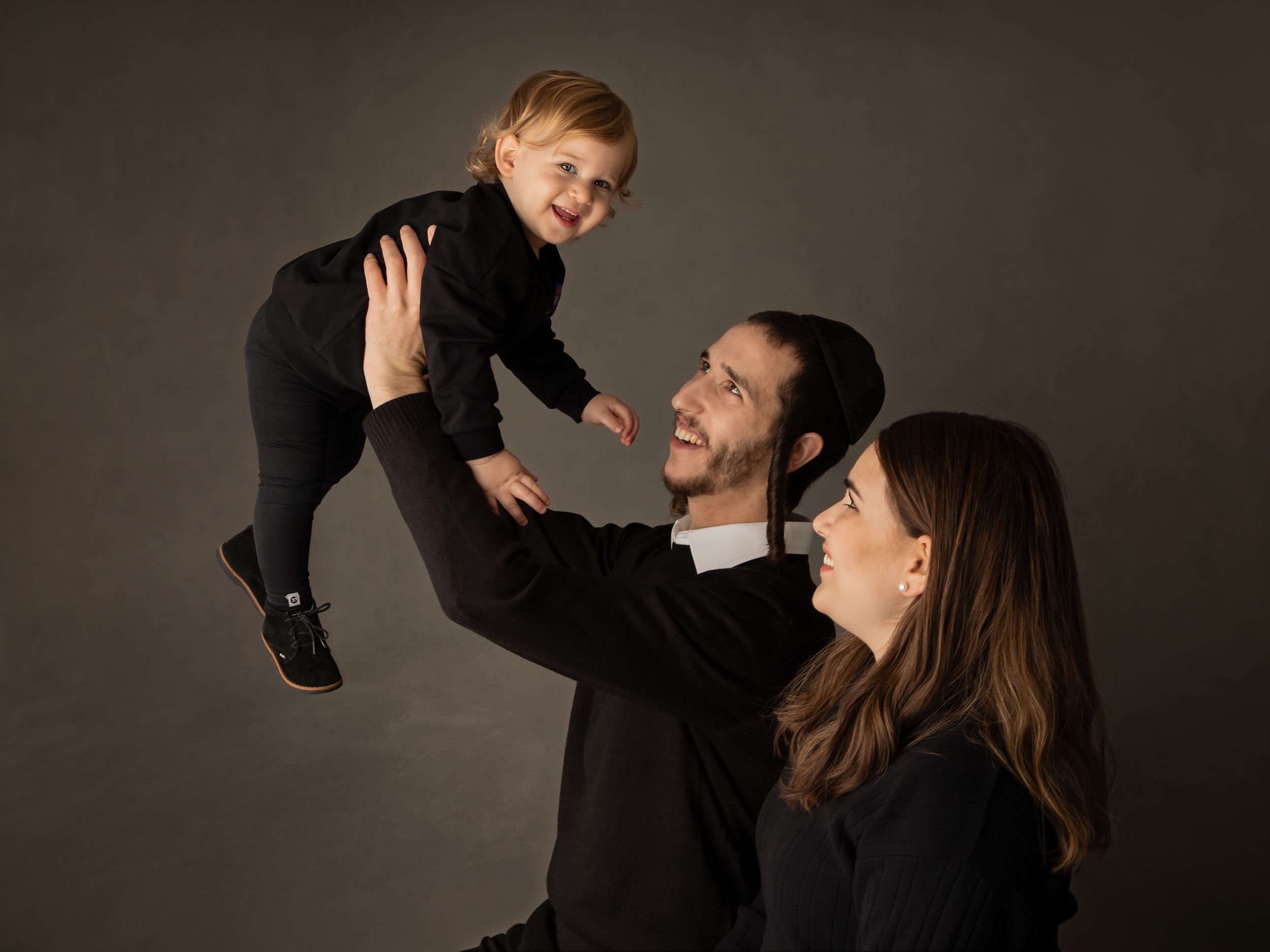 A family in dark clothing posing together against a brown backdrop for a portrait session.