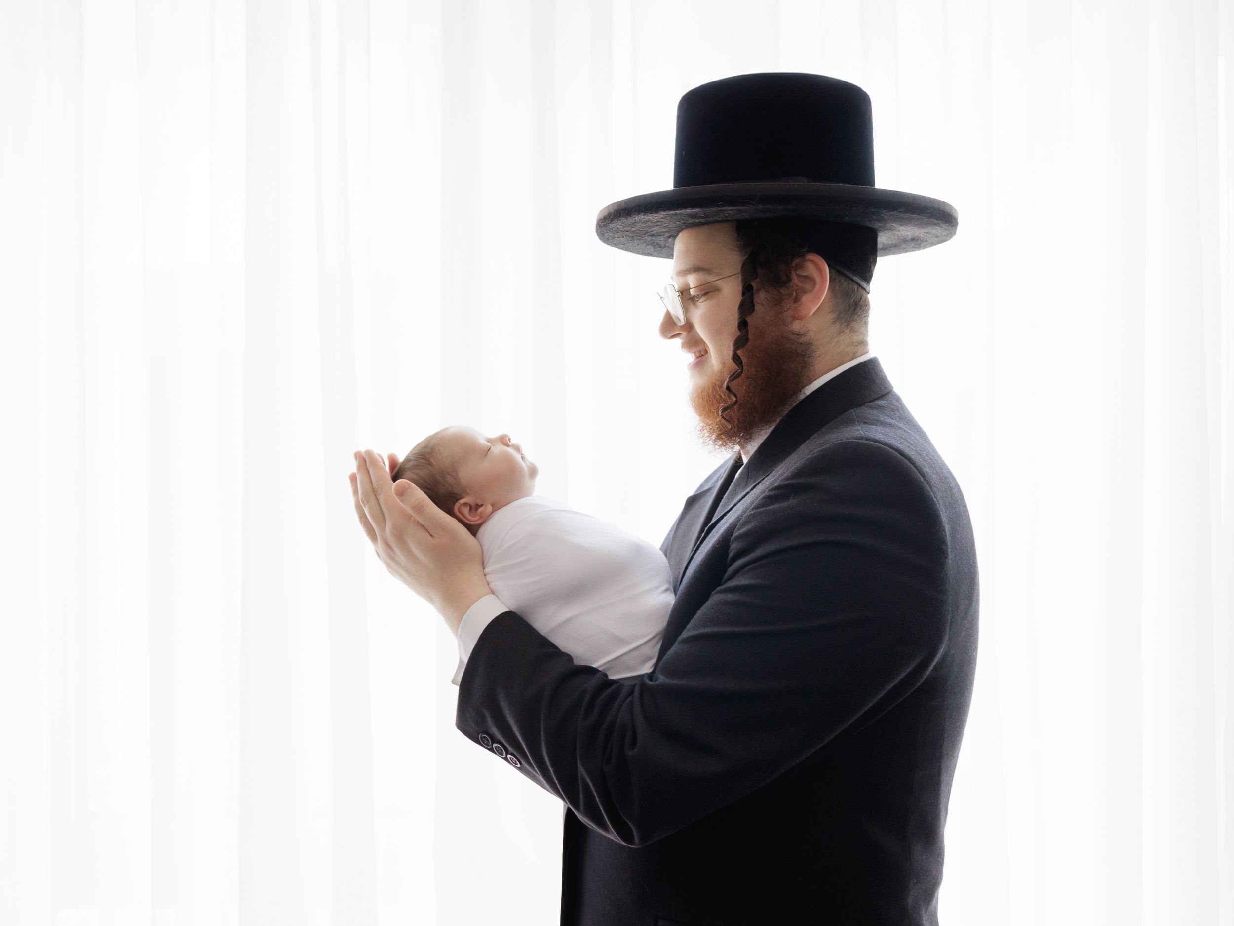 Side view of person in traditional black suit and hat tenderly holding a swaddled infant against white background.