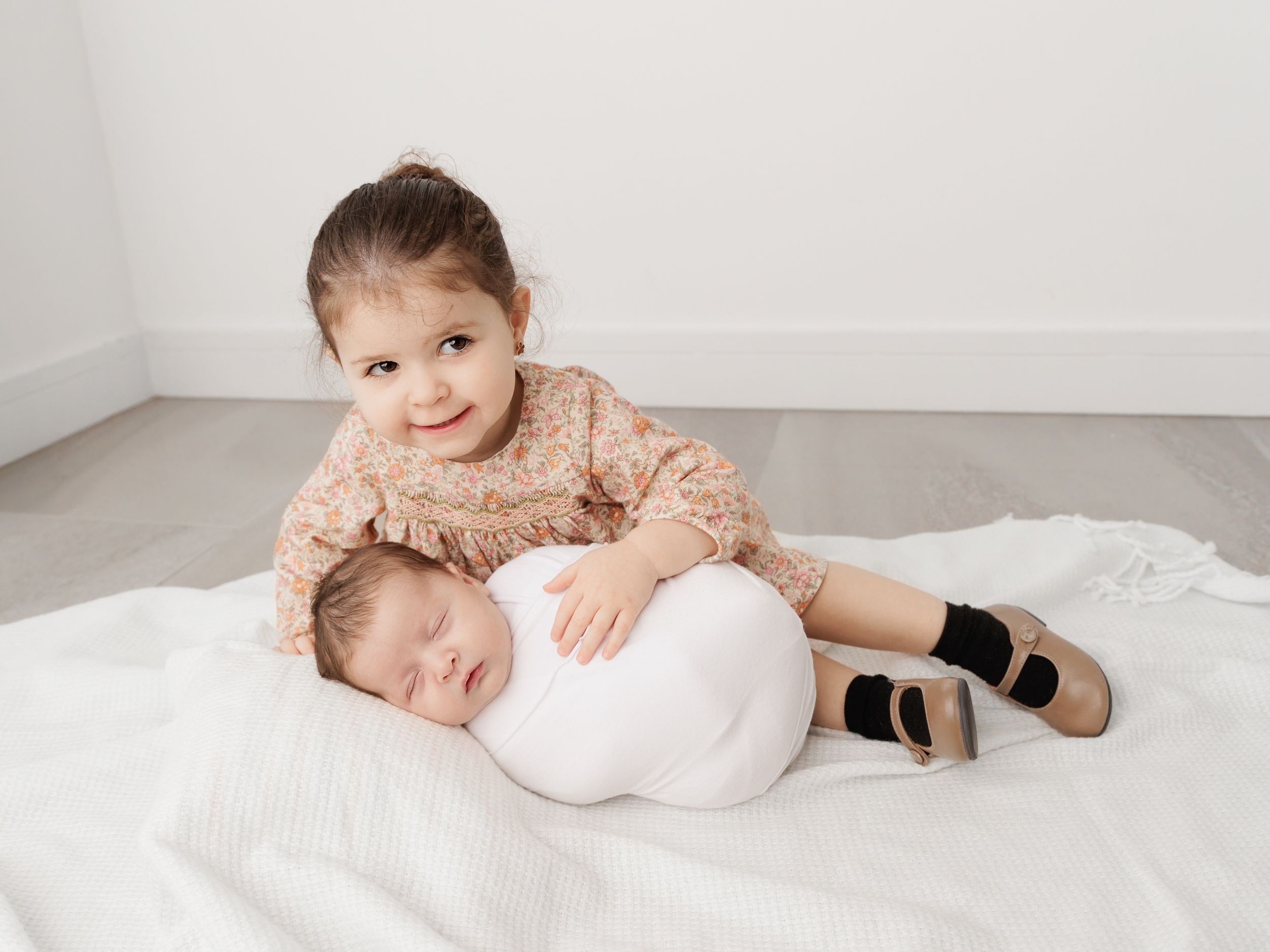 Two young children on a white blanket, with one laying down while the other sits beside wearing a peach lace dress and black shoes.