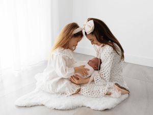 Two girls in white dresses sharing a loving moment with a newborn on a cozy white blanket.