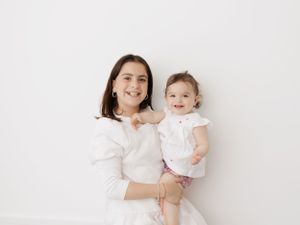 Mother and child in matching white outfits pose against white wall.