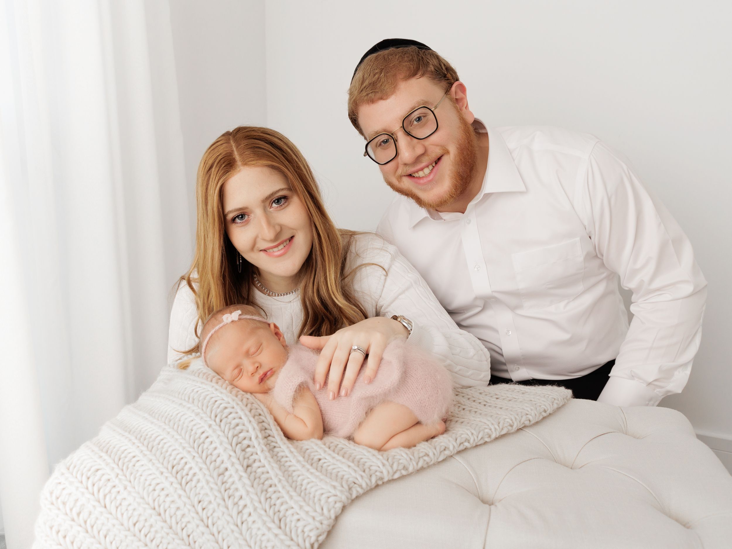 A sleeping newborn baby wearing pink lies on a white knit blanket while parents in white shirts look on.
