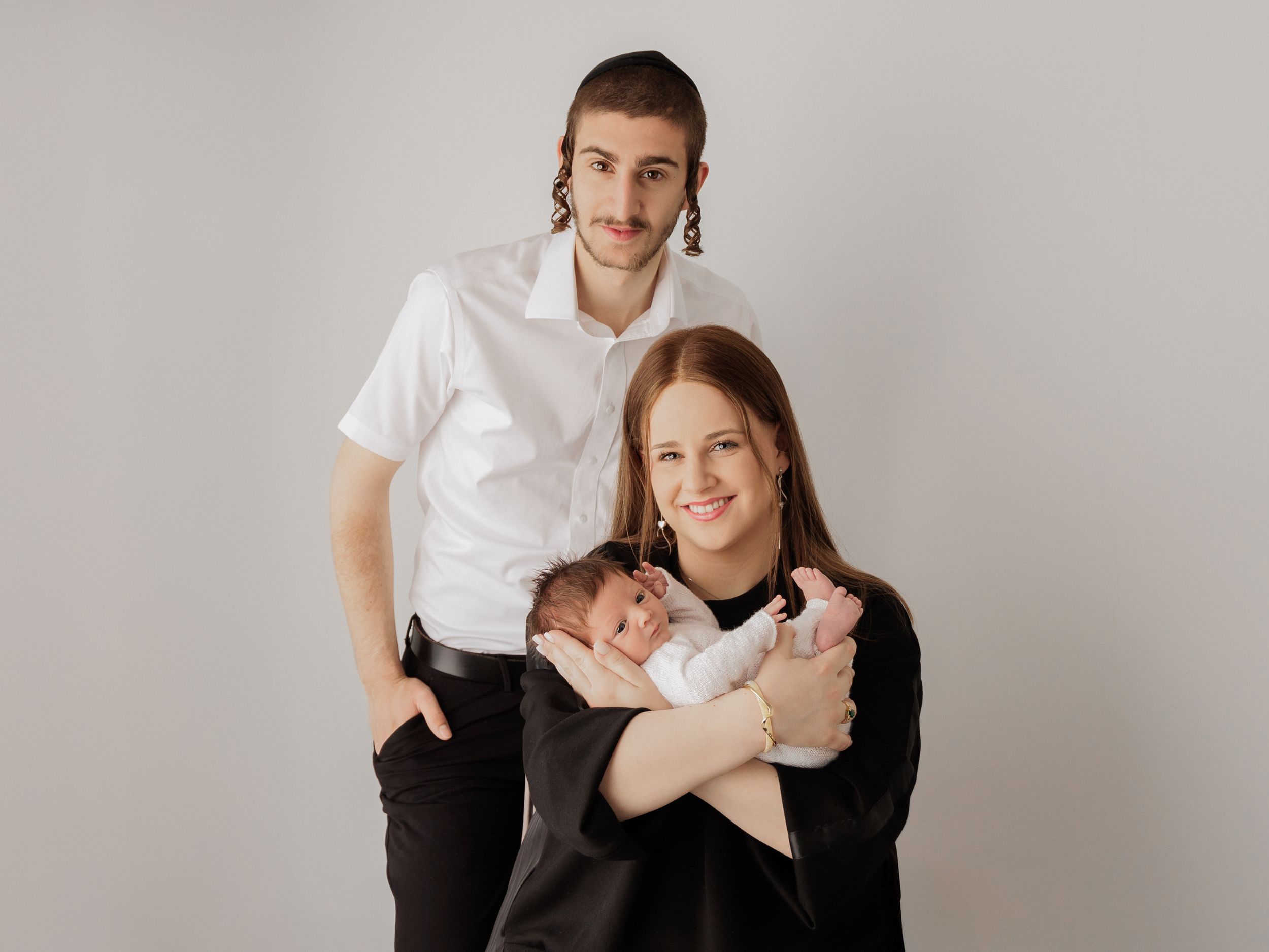 A couple wearing black and white outfits holds their newborn baby against a plain gray studio background.