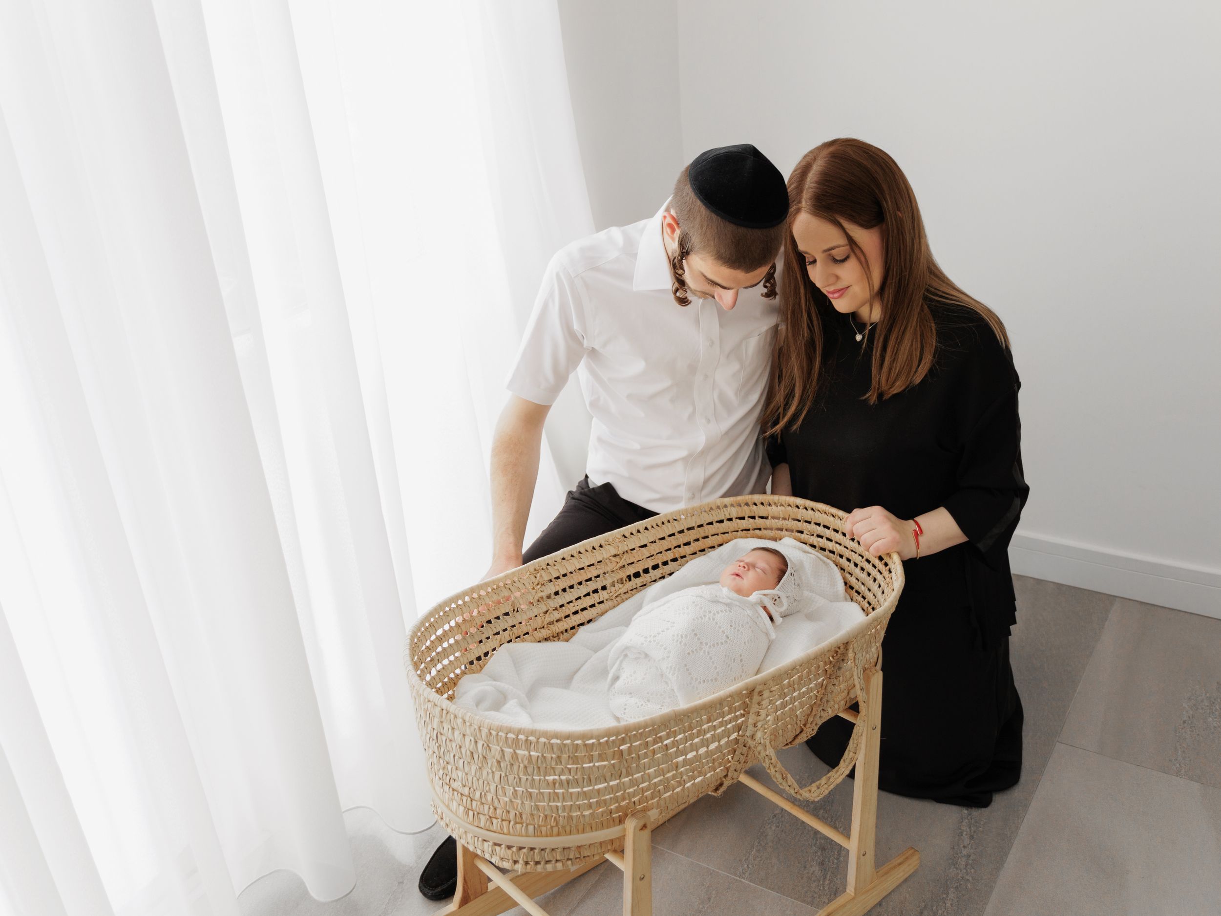 A couple stands over a woven rattan baby bassinet in a bright minimalist room with white walls and curtains.