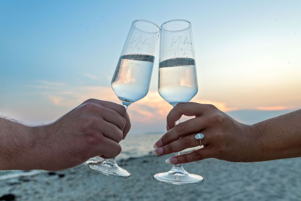 Two hands clink wine glasses together against a sunset beach backdrop creating a romantic seaside moment.
