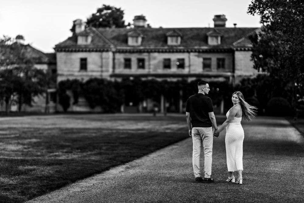 A couple stands together on a path leading to a grand Georgian-style mansion in black and white photography.