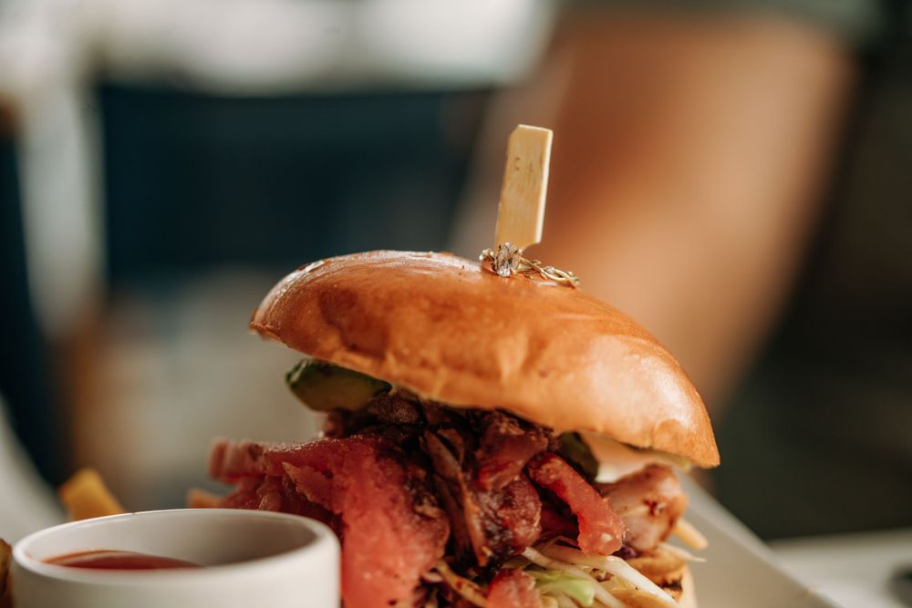 A gourmet sandwich with piled high deli meats and a golden toasted bun served alongside a dipping sauce.