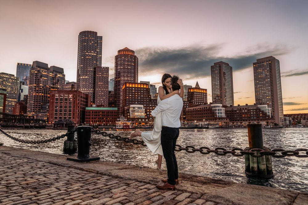 A person stands on a harbor pier during sunset with Boston's stunning downtown skyline in the background.