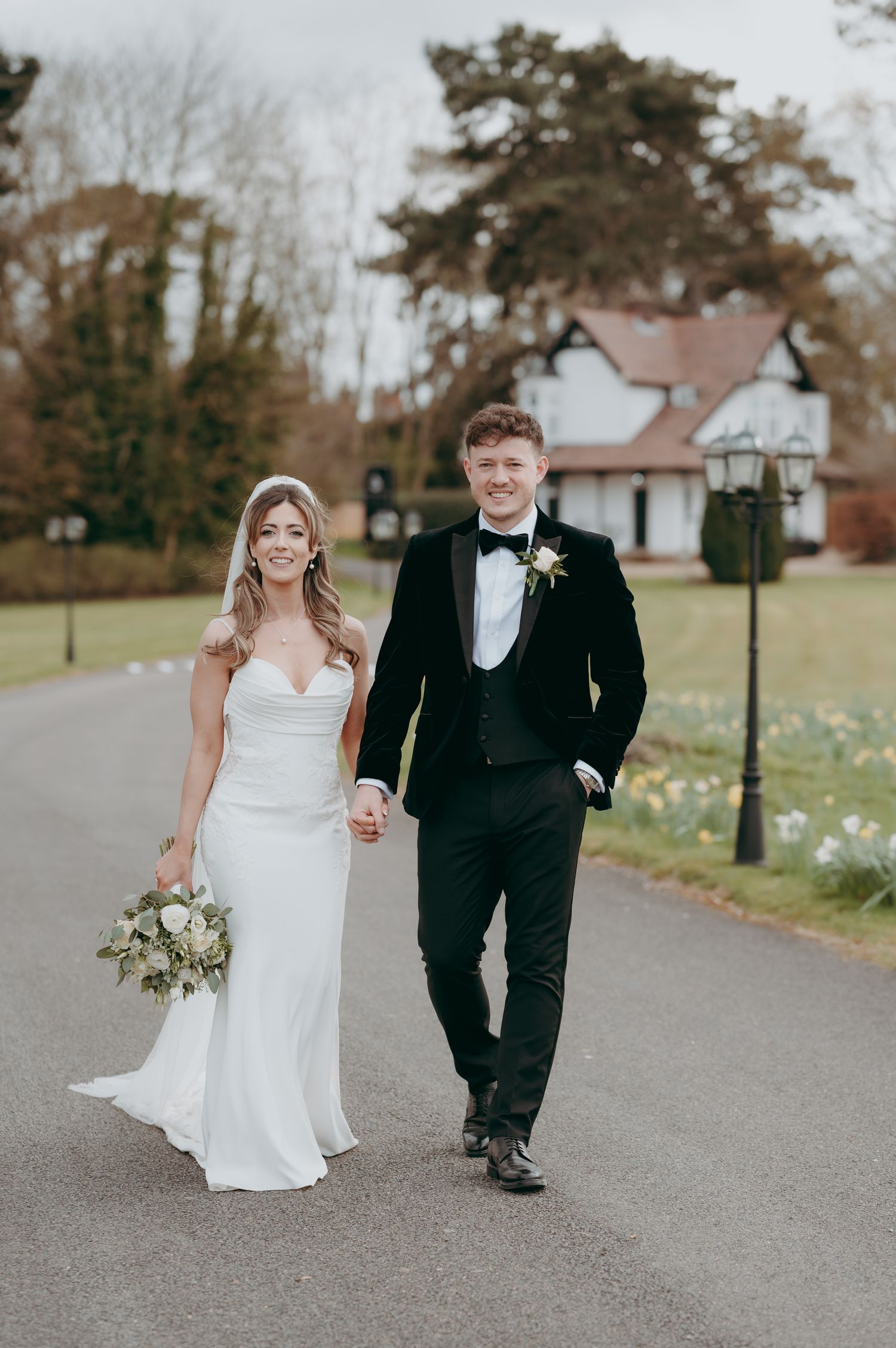 Couple in wedding attire walks hand in hand down a tree-lined path with daffodils in spring. Swynford Manor