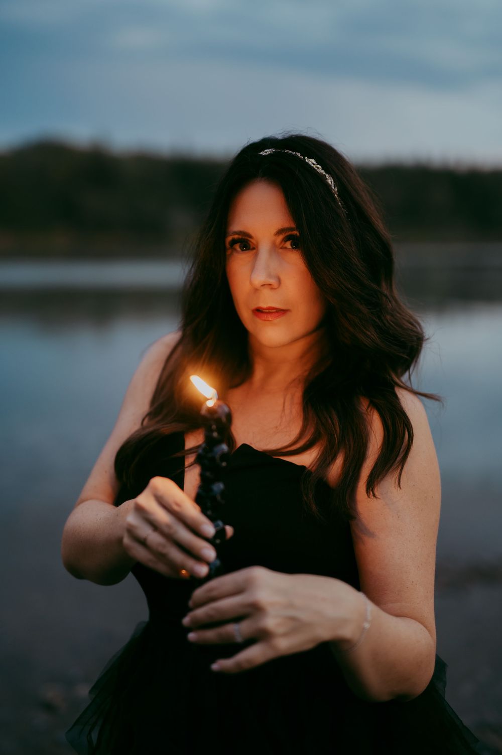 Portrait of a person in a black dress holding a lit candle near a lake at dusk.