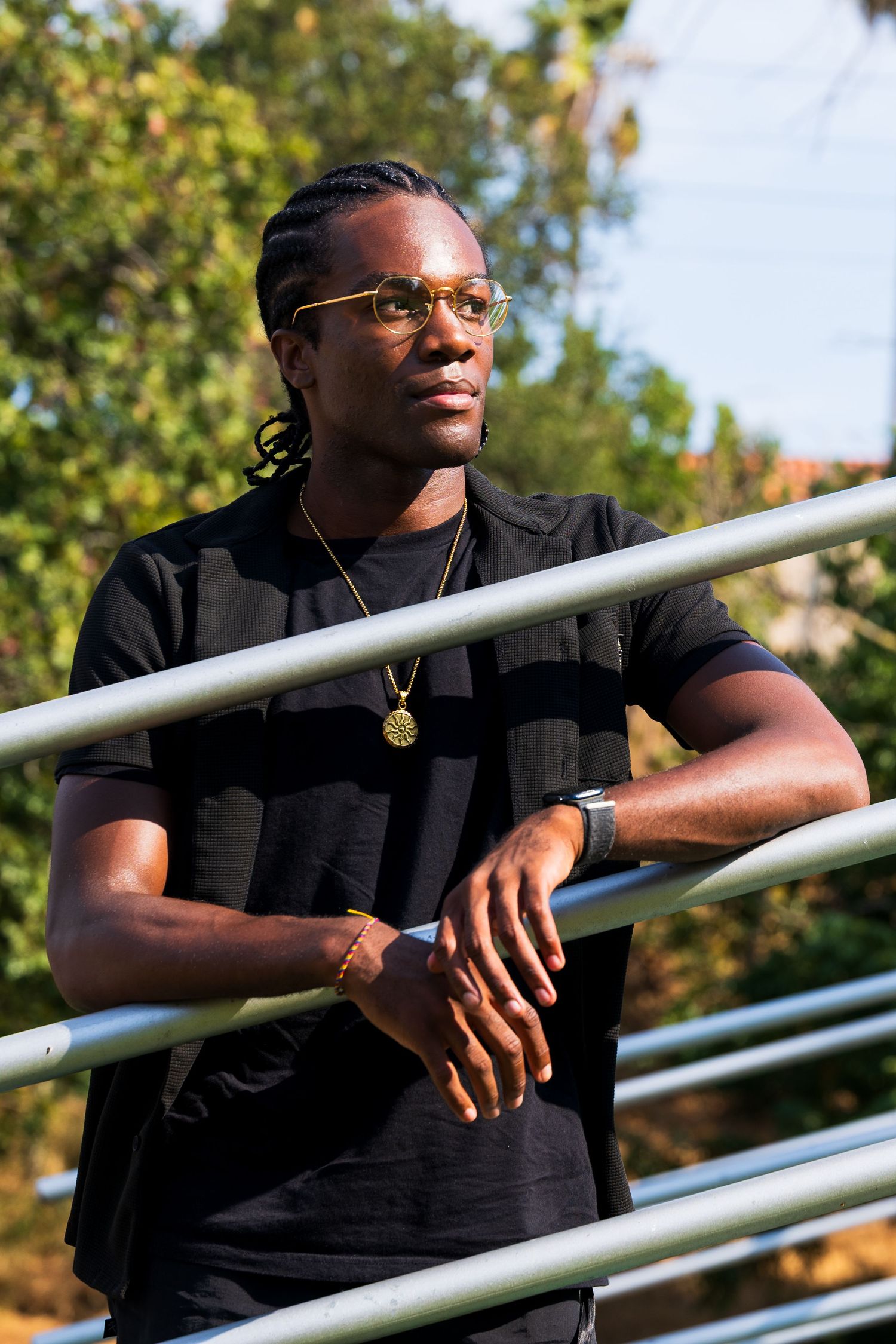 An outdoor portrait of a black man, Mwendwa Rwakatare, in black attire leaning thoughtfully against metal railings at golden hour.