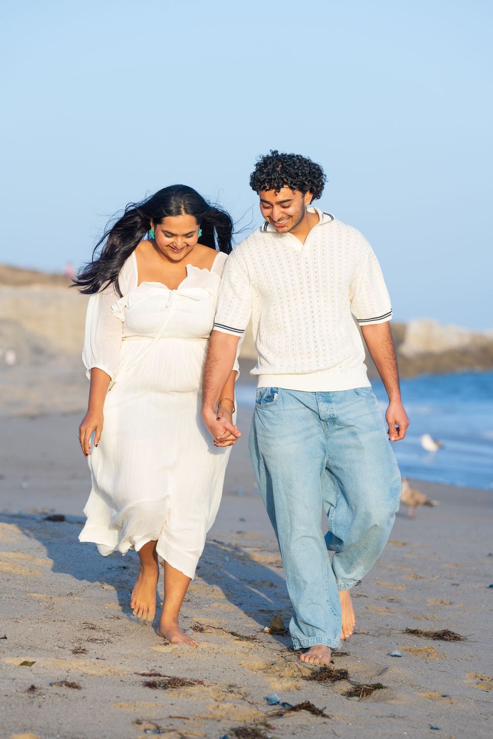 Couple portraits of a man and woman holding hands and walking on the beach sand in LA county. They both wear white tops.