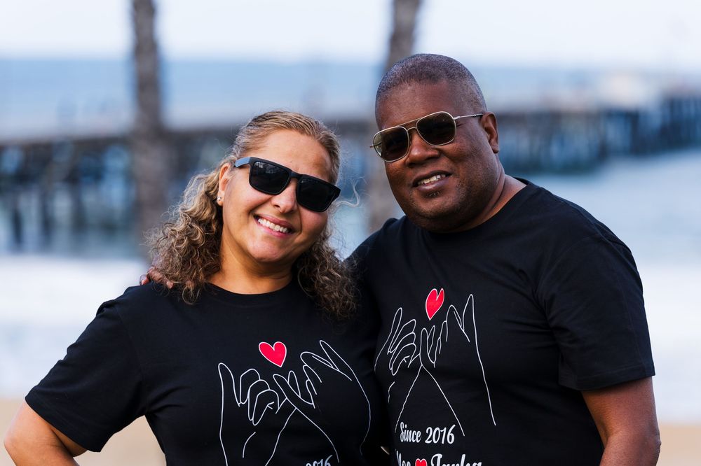 A mixed race couple wears matching tees for their 10yr anniversary while posing for photos at the beach in front of the pier.