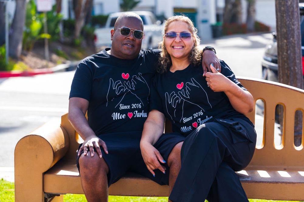 A mixed race couple sits on a bench at a park. The man puts his hand over the woman's shoulder and she holds his hand