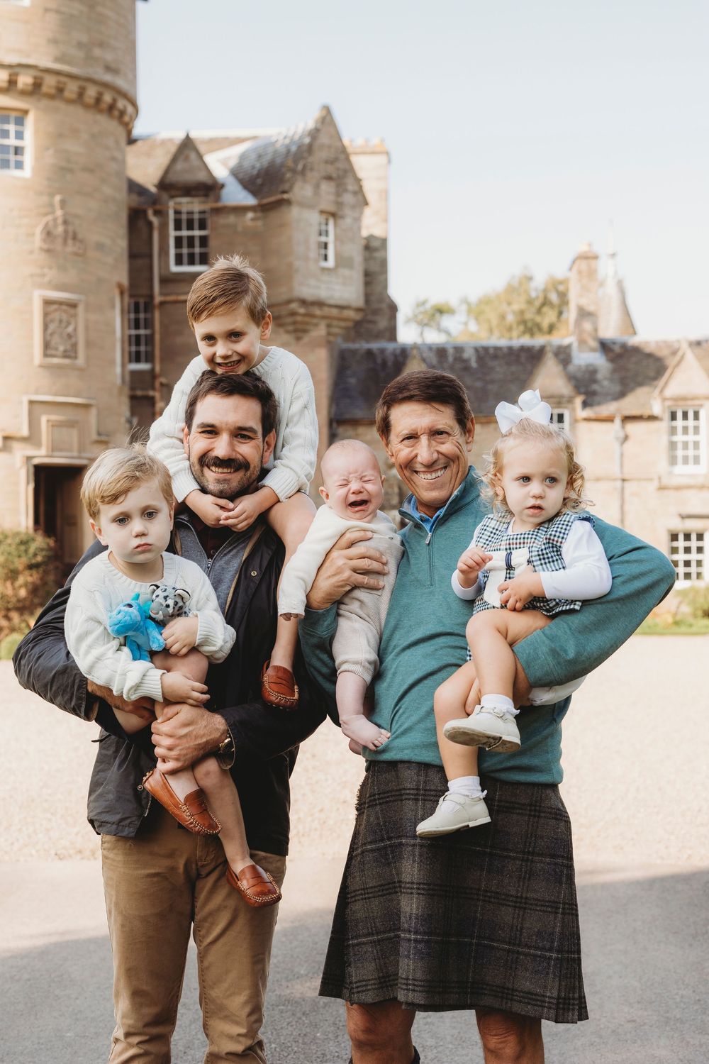 Two men holding four young children pose smiling outside a historic Scottish castle, one man wearing a traditional tartan kilt.