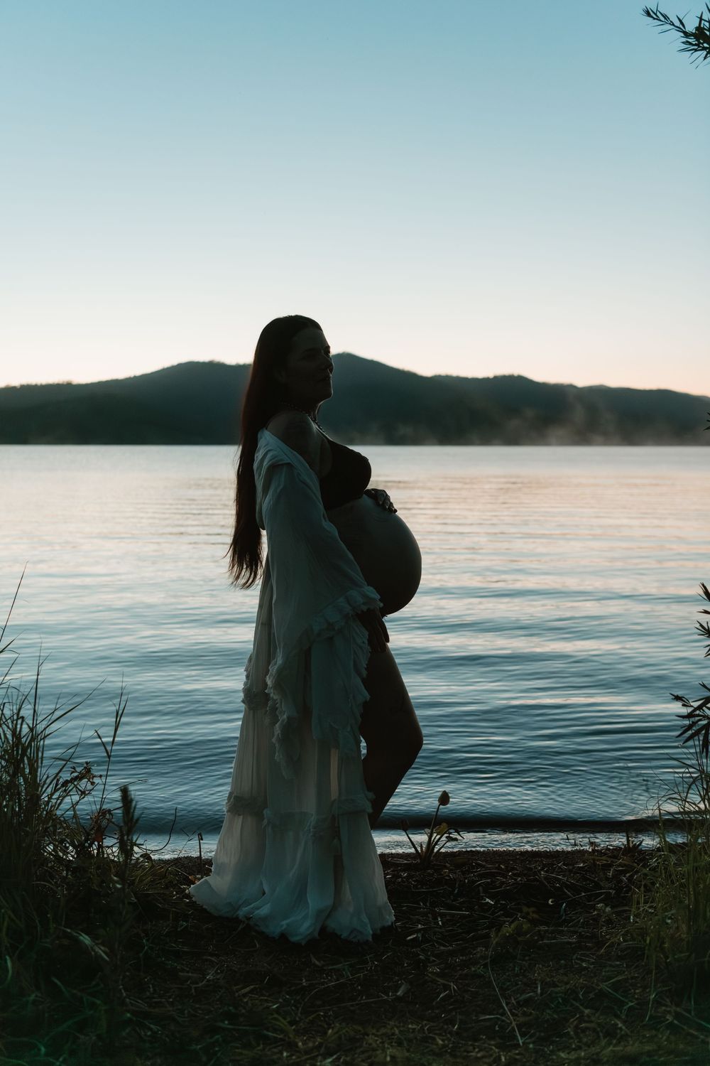 Pregnant woman in white dress standing by a serene lake at dusk with misty mountains in the background.