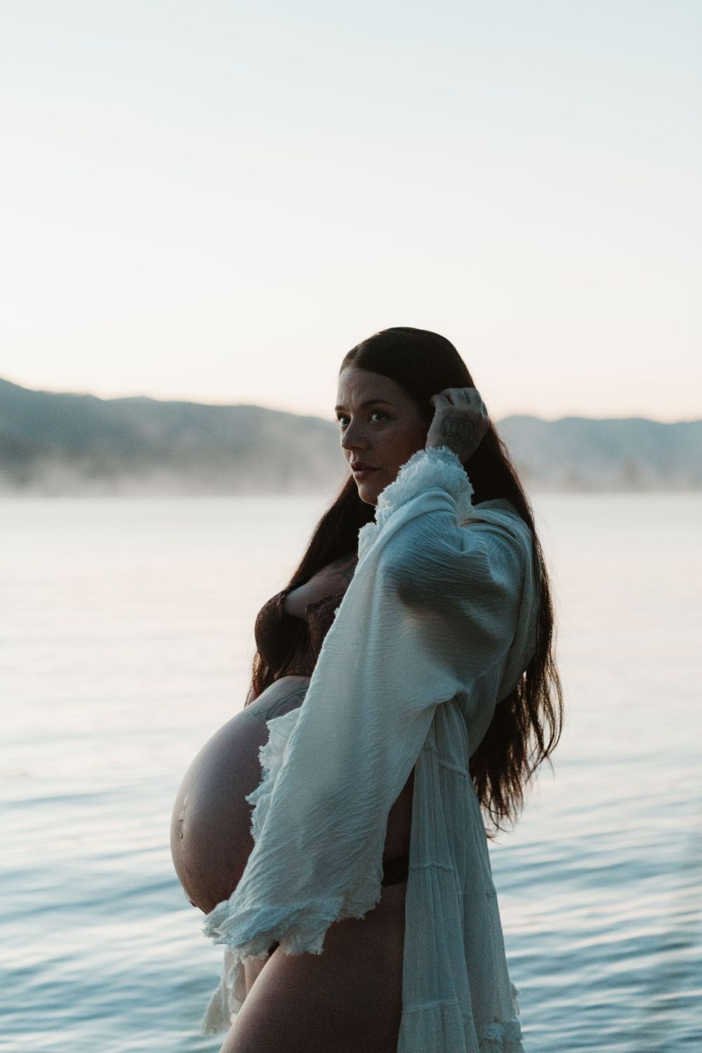 Pregnant woman in white wrap standing by a serene lake at dusk with misty mountains in the background.