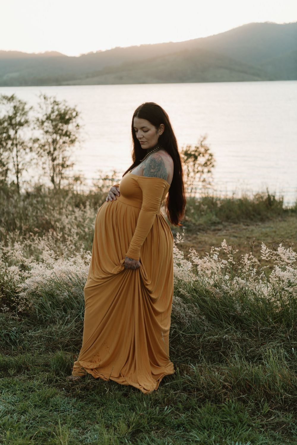 Pregnant woman in golden yellow off-shoulder maxi dress cradling baby bump outdoors near lake at sunset.