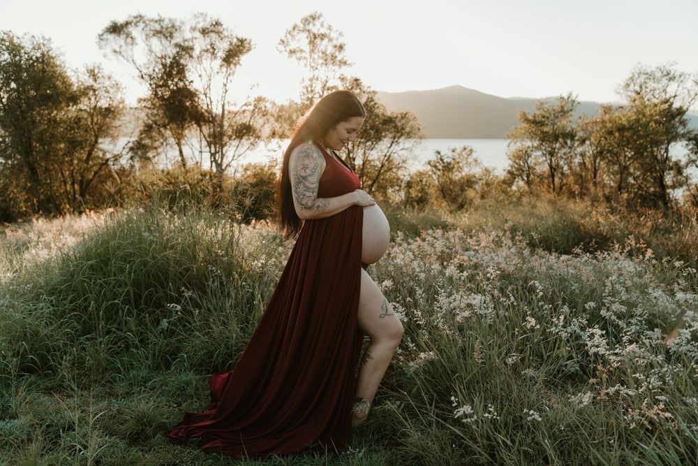Pregnant woman in burgundy gown standing in golden meadow at sunset near lake.