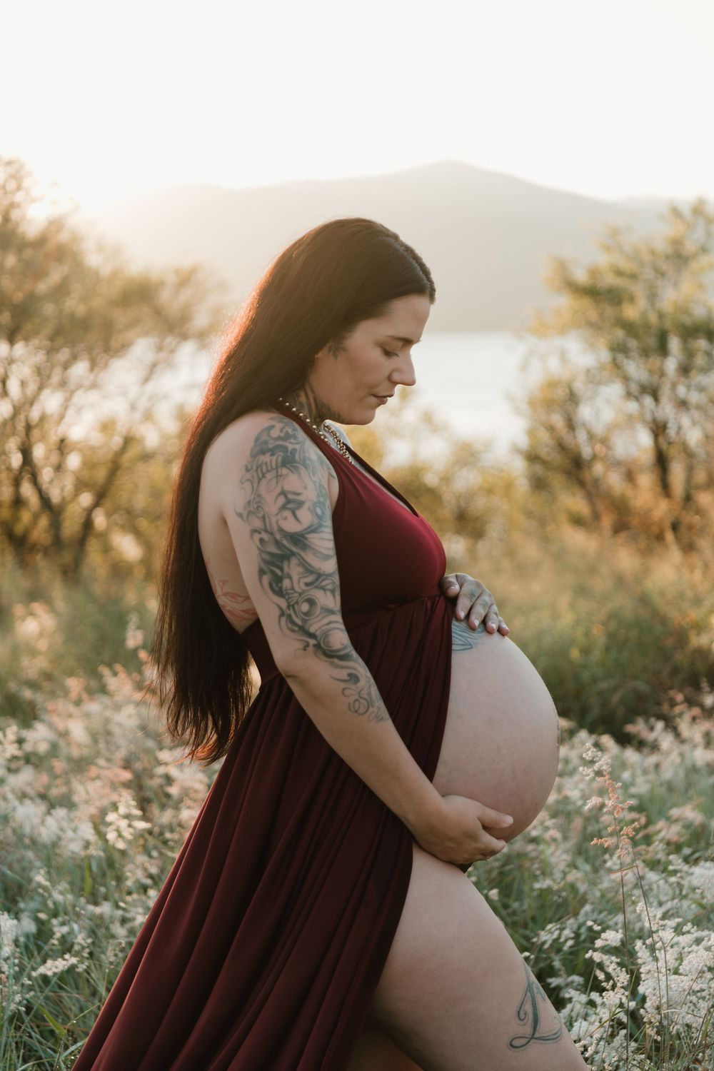 Pregnant woman in burgundy dress holding baby bump at golden hour in a field with tattoos on her arm.