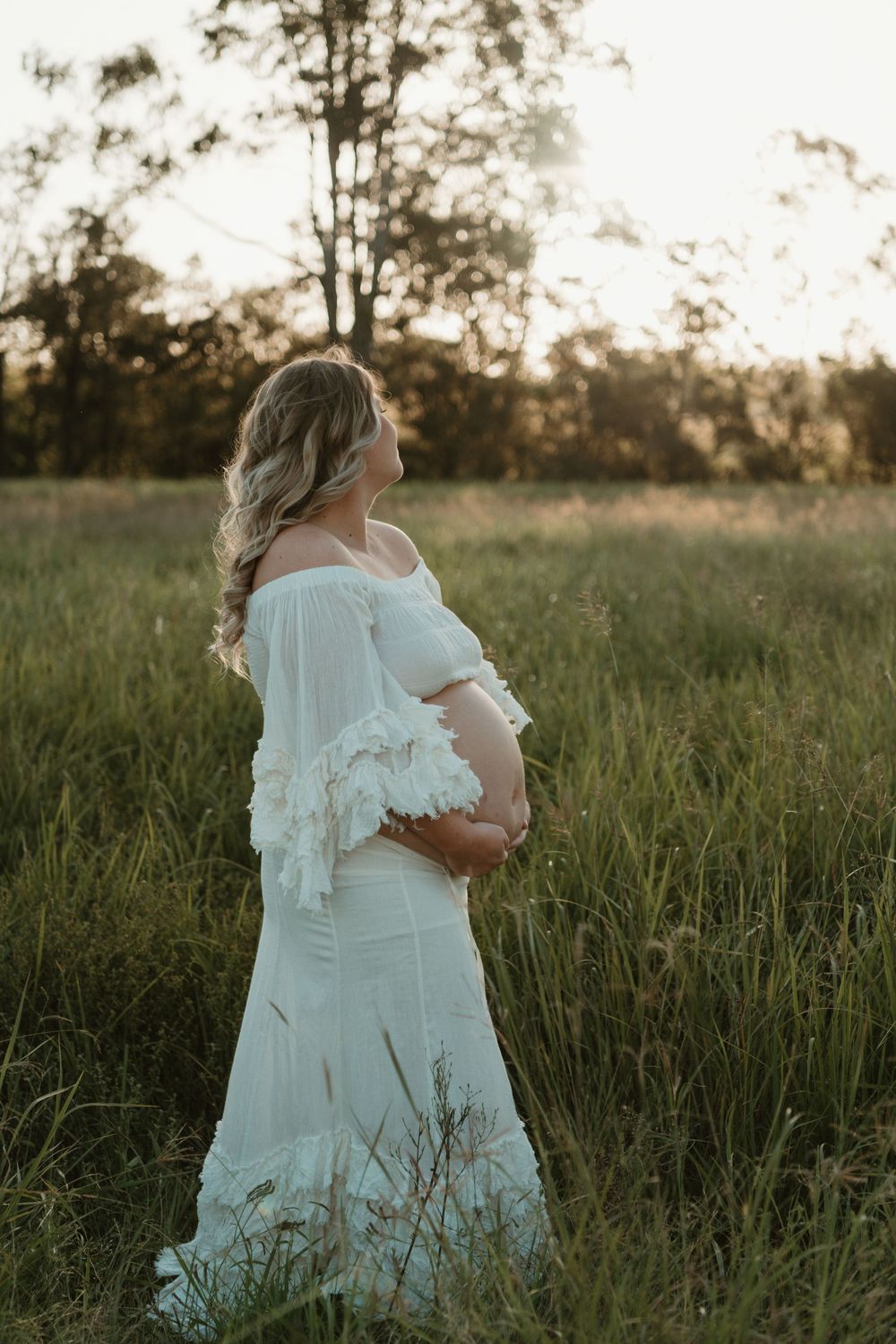 Pregnant woman in white off-shoulder dress holding baby bump in golden hour field.