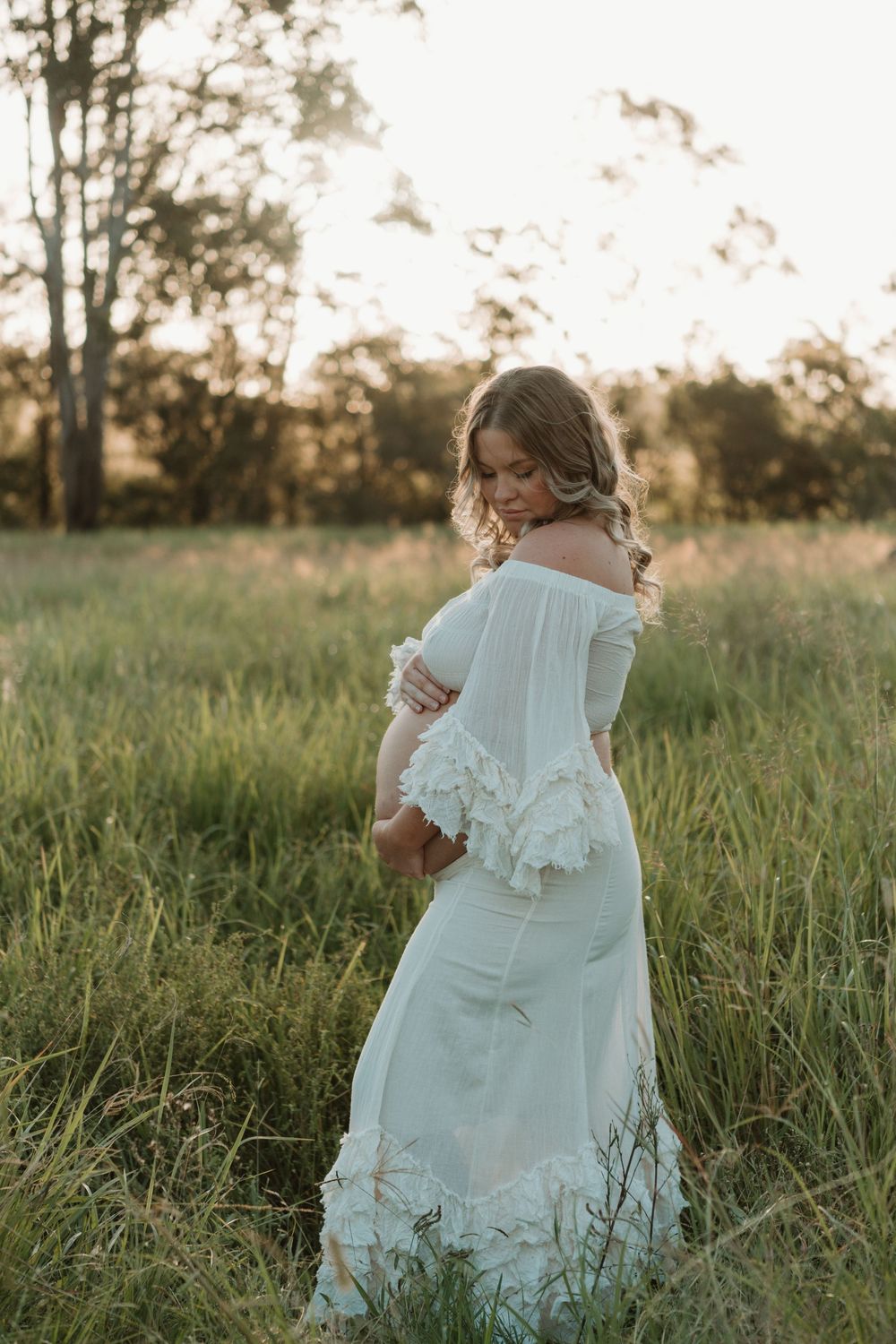 Pregnant woman in white off-shoulder ruffle dress standing in green field at golden hour sunset.